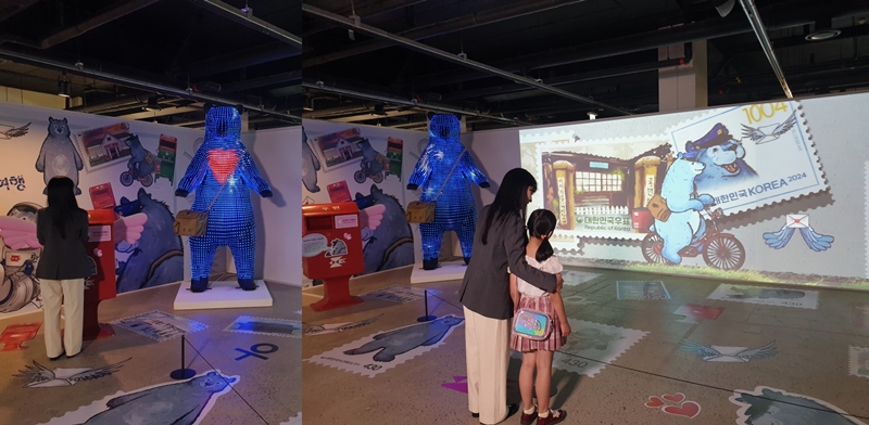 El 18 de julio, unas visitantes de la Exposición Filatélica de Corea 2024 observan una obra del artista Lee Jae-hyung que presenta un oso azul viajando a través de 140 años de historia de los sellos postales en una galería en la isla Nodeulseom, en el distrito de Yongsan-gu, Seúl. | Koh Hyunjeong