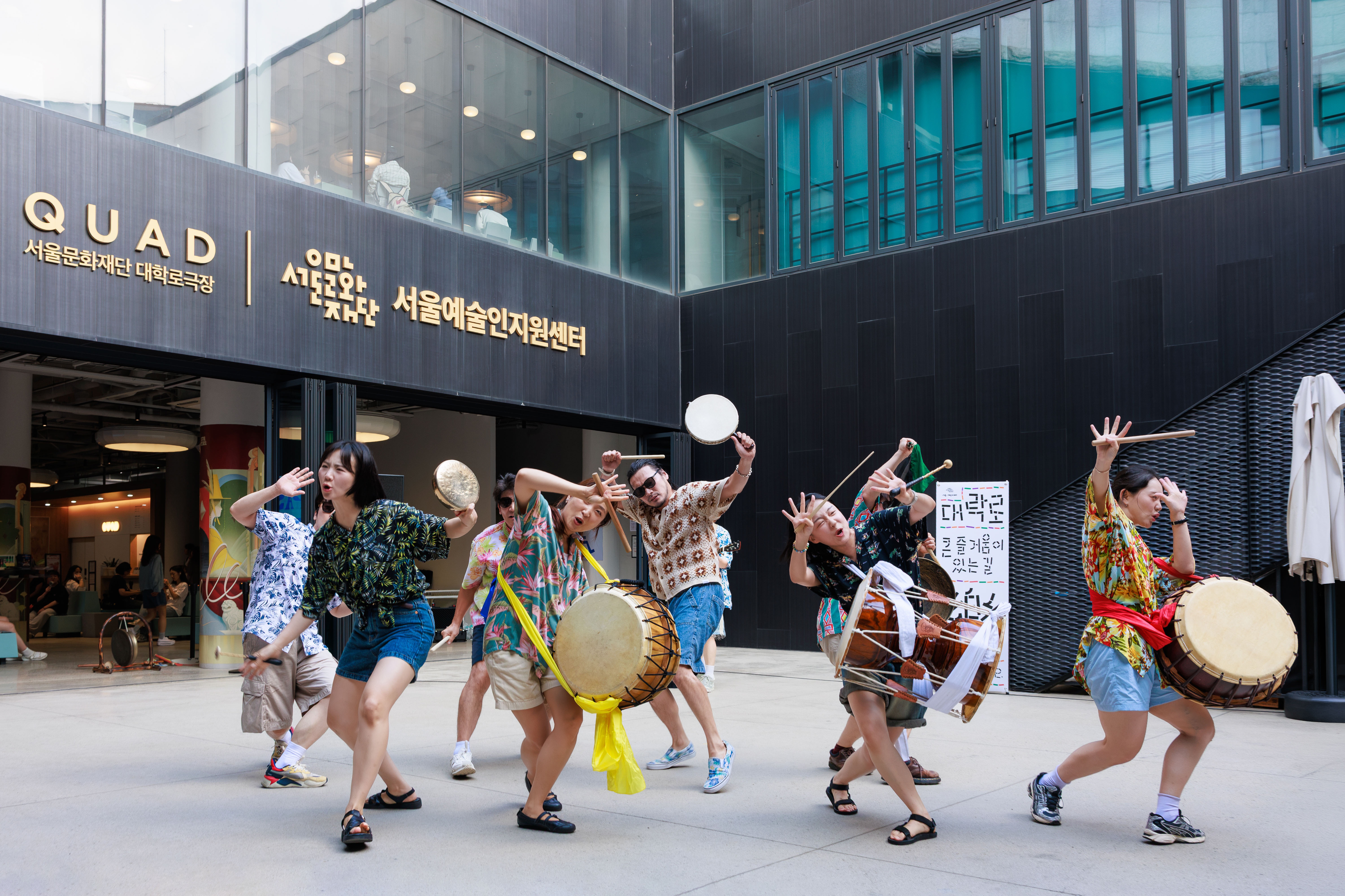 La imagen muestra al elenco de la obra de teatro '¡El teatro continúa!' realizando una representación al aire libre al frente de la Fundación para las Artes y la Cultura de Seúl. | Fundación para las Artes y la Cultura de Seúl