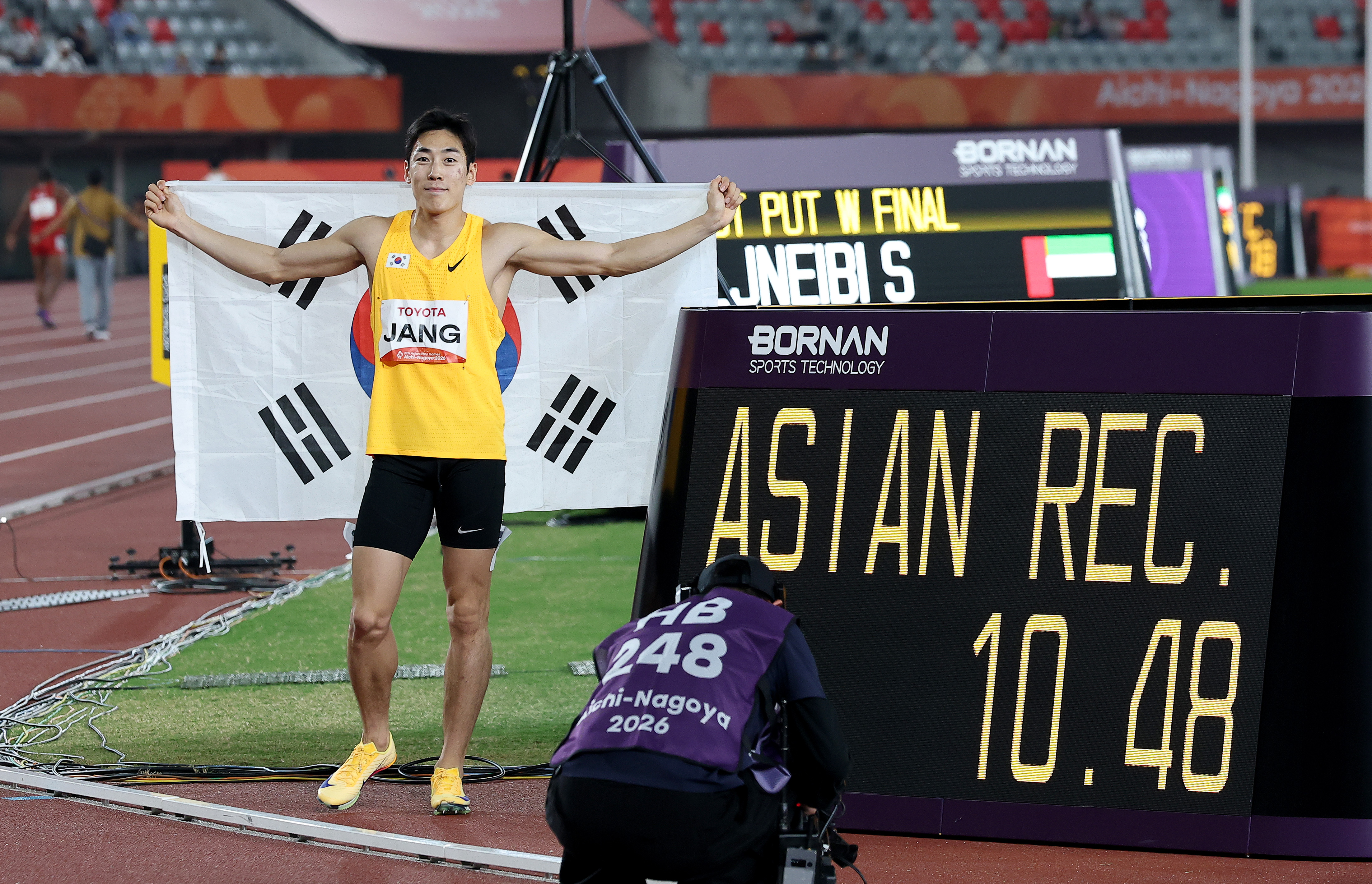 El velocista coreano Jang Minho posa junto al marcador oficial tras adjudicarse la medalla de oro con una nueva marca continental de 10,48 segundos en la final masculina de los 100 metros en la clase T13 de los Juegos Para Asiáticos de Aichi-Nagoya 2026, celebrada el 20 de octubre en el Estadio de Atletismo Mizuho en Nagoya, Japón.