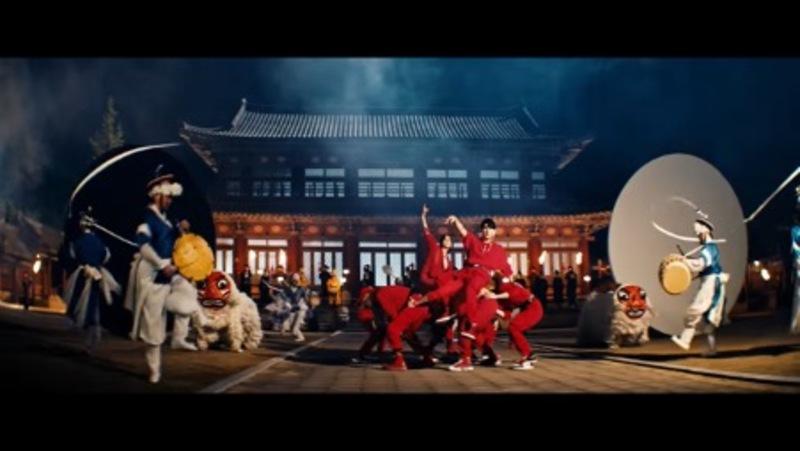 Integrantes de Stray Kids de color rojo y junto a ellos bailarines de Samulnori con vestimenta azul y blanca. En el fond