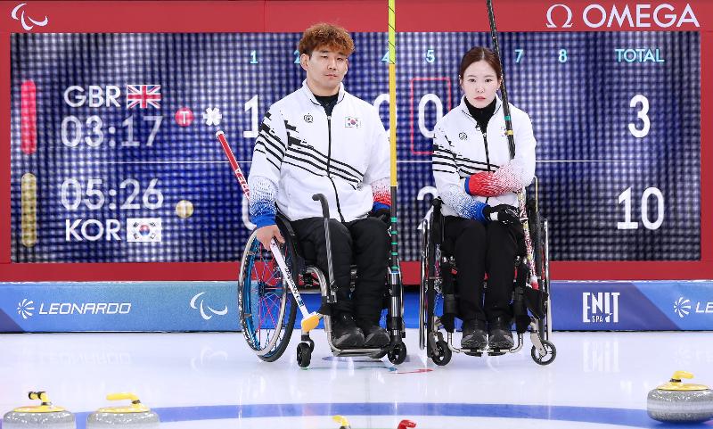El 6 de marzo (hora local), la pareja de dobles mixtos de curling en silla de ruedas, conformada por Lee Yongsuk (izda.) y Baek Hyejin, observa las piedras durante el encuentro contra el Reino Unido. Al fondo, se aprecia el marcador que refleja la amplia ventaja de la delegación coreana, sellando una victoria contundente frente al equipo británico. | Comité Paralímpico de Corea 